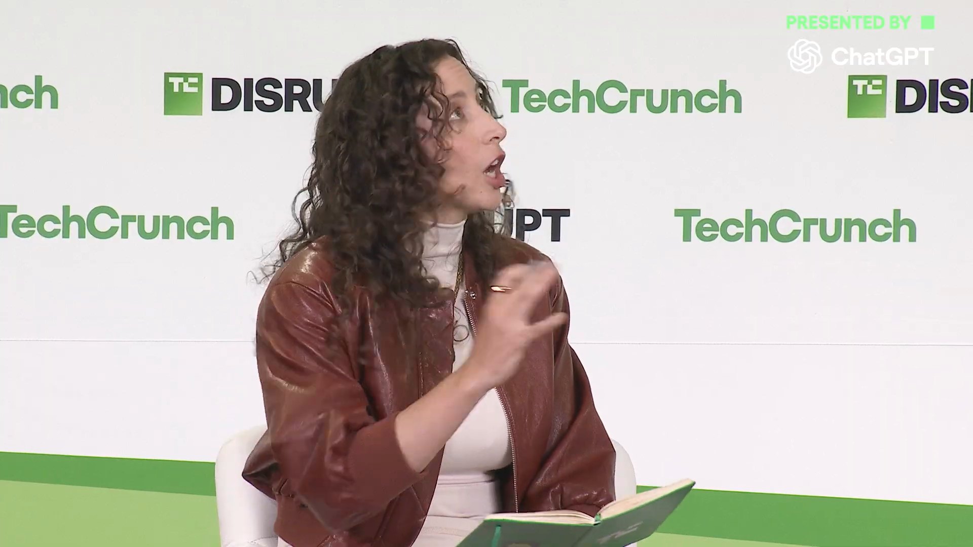 TechCrunch 資深記者 Rebecca Bellan 手拿綠色筆記本，身體前傾、單手比劃地向 Masad 提問，背景是綠白相間的 TechCrunch 布幕