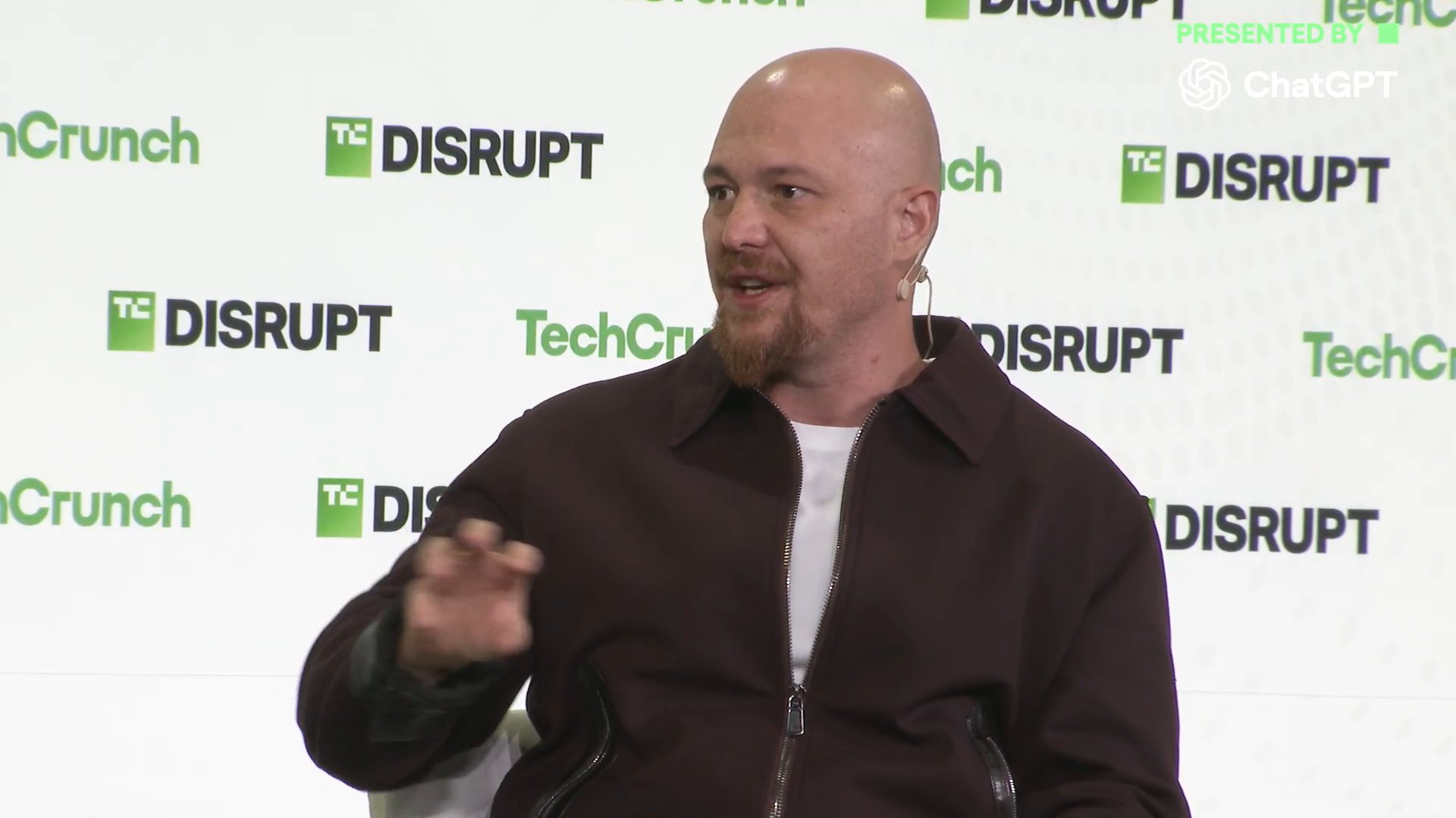 Amjad Masad 轉向主持人、單手比劃解釋，背景是 Disrupt 與 TechCrunch 的綠白布幕