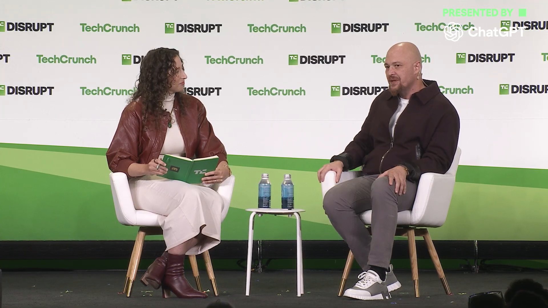 Disrupt 2026 舞台上，Replit 執行長 Amjad Masad 坐在白色扶手椅上，與手拿綠色筆記本的 TechCrunch 資深記者 Rebecca Bellan 面對面對談，背景是 TechCrunch 與 Disrupt 的綠白相間布幕